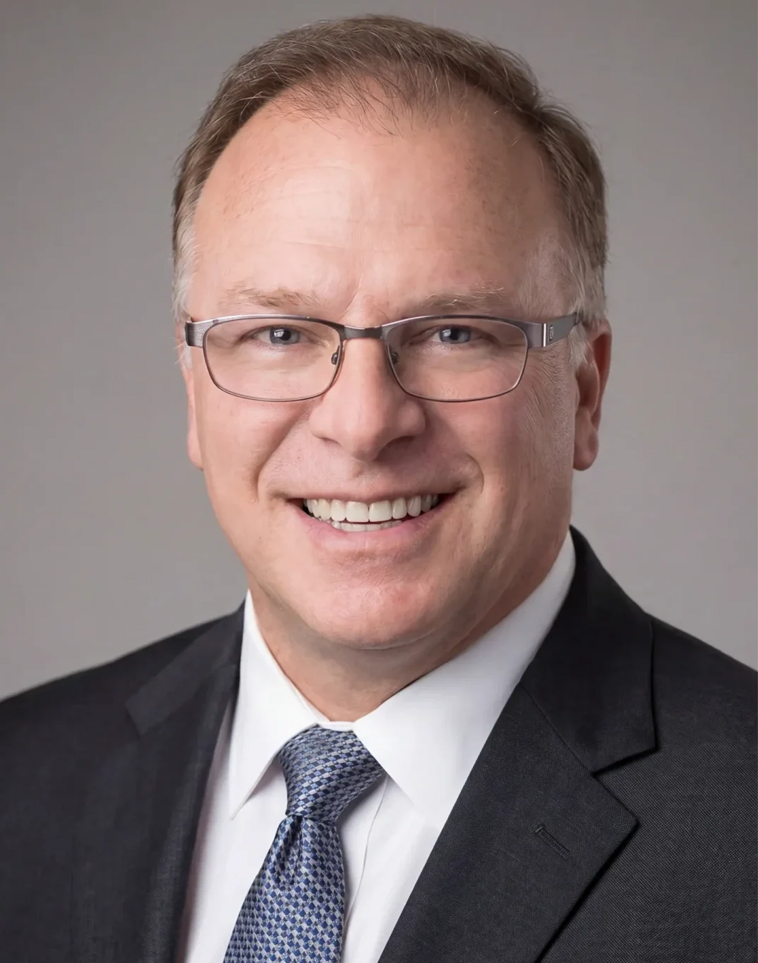 Close-up of a smiling man wearing glasses and a dark suit, white shirt, and patterned tie, with a gray background.